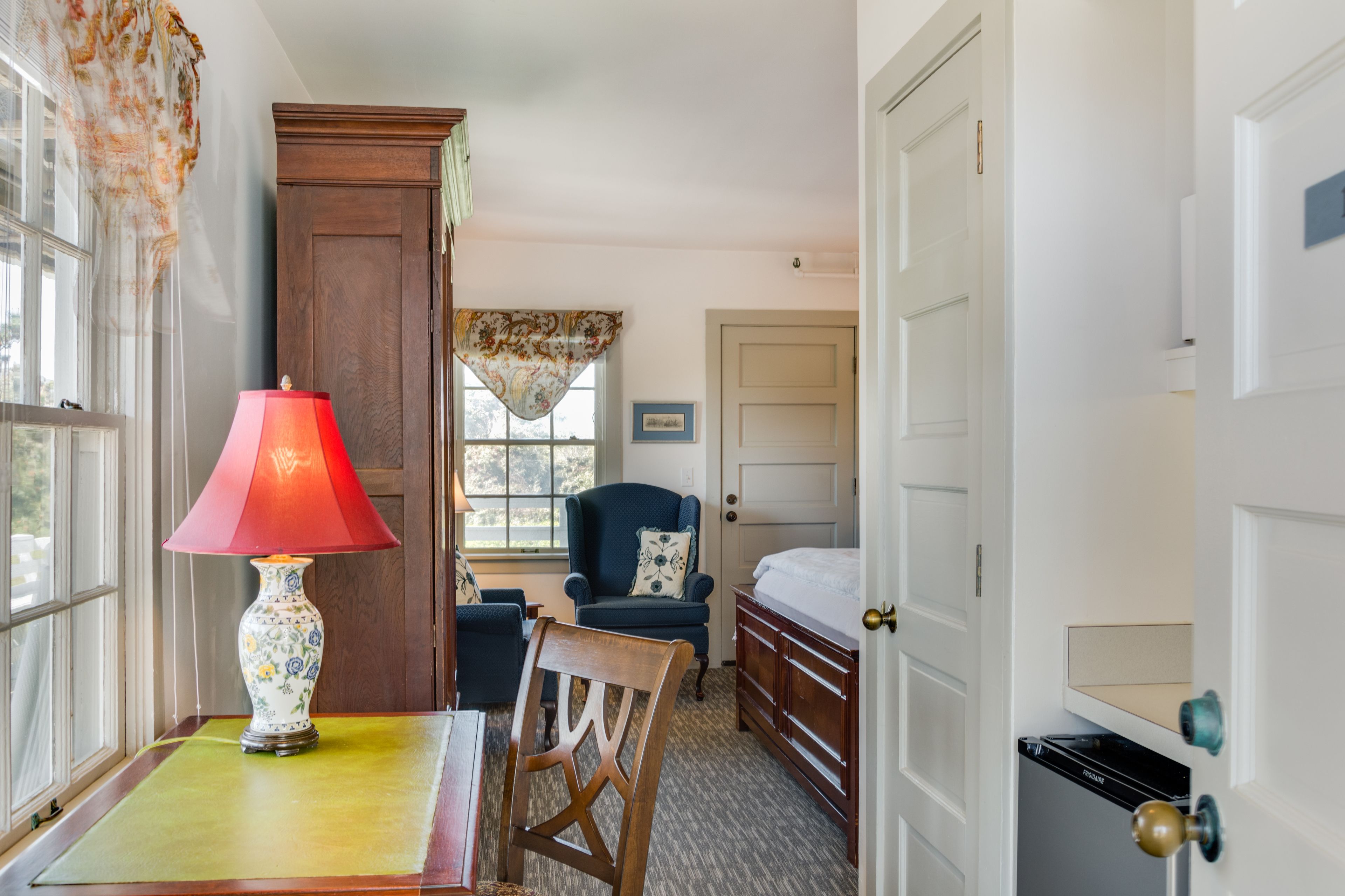 A room with a wooden chair, a table with a red lamp, a bed, and a door.
