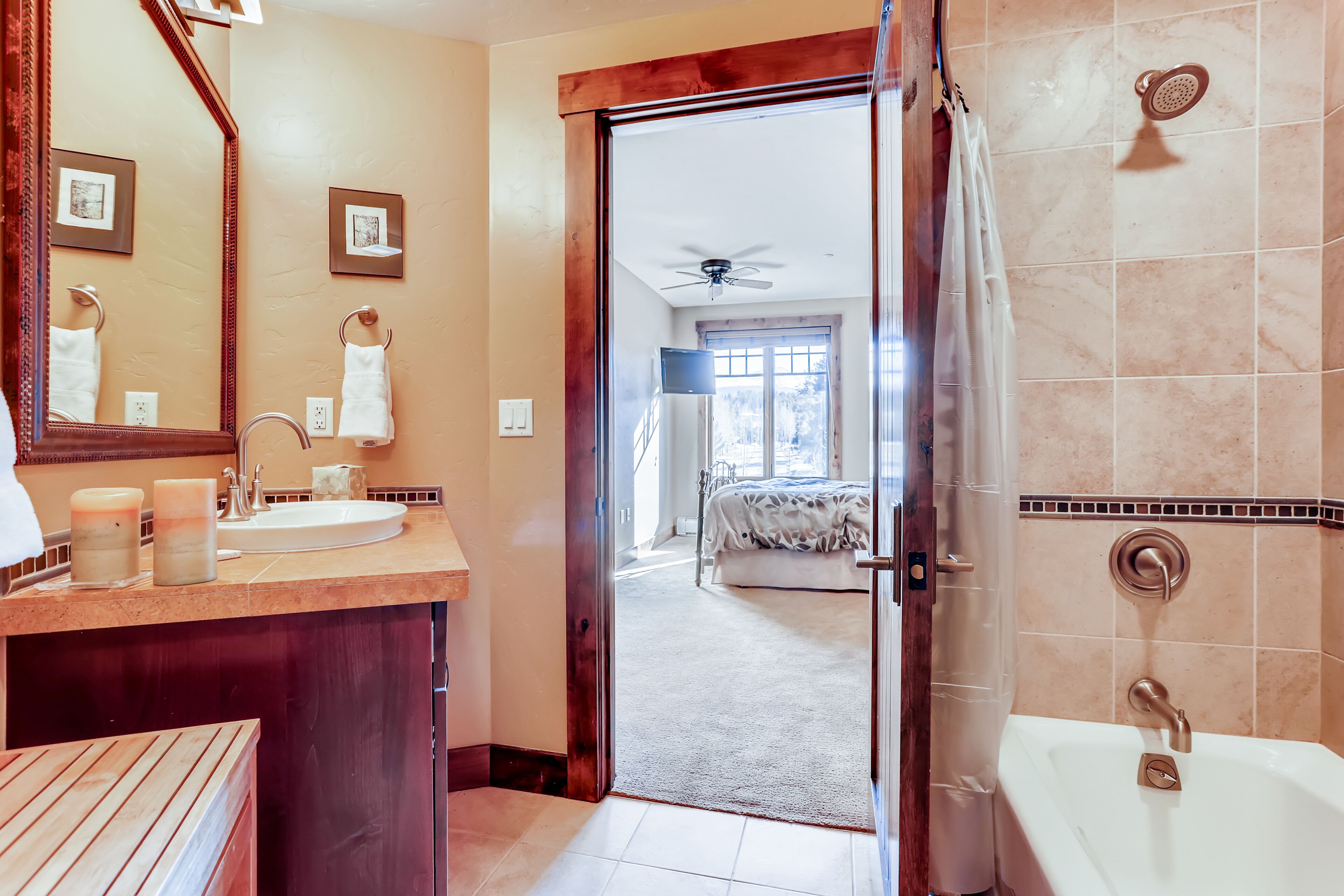Ein Badezimmer mit Waschbecken, Spiegel und Dusche. Eine Tür führt in einen Schlafbereich mit Bett und Fenster.
