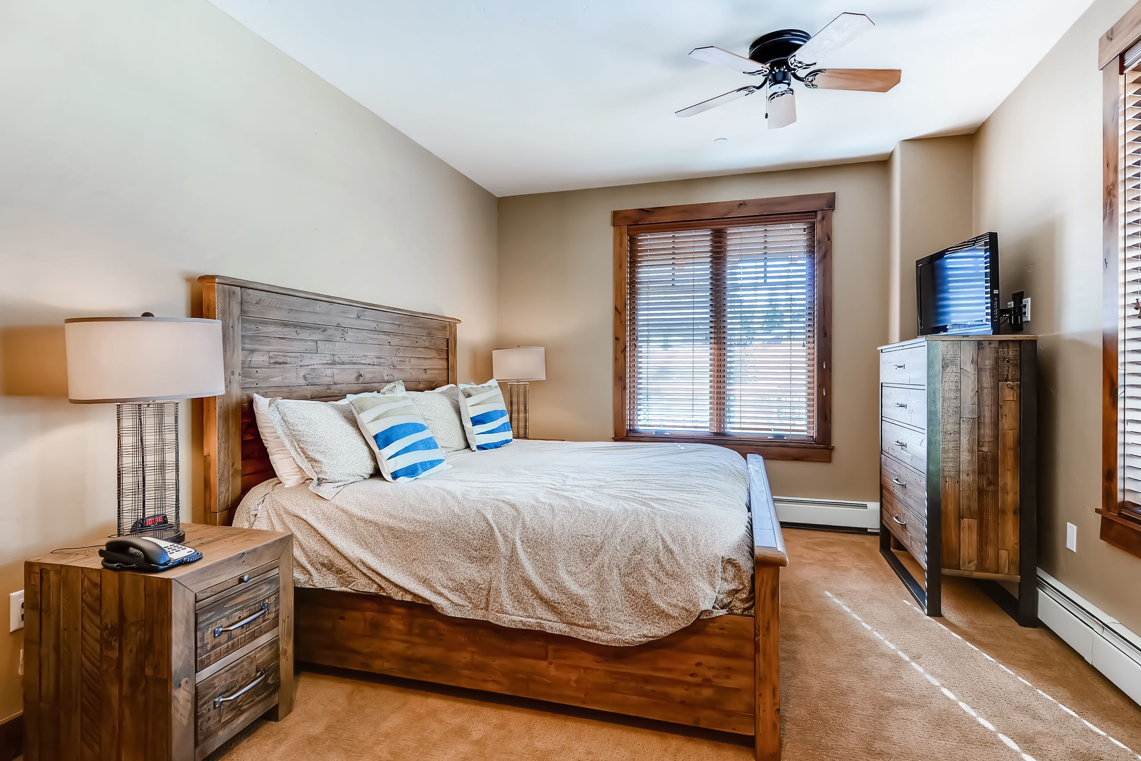A bedroom with a wooden bed, two bedside lamps, a nightstand, a dresser, and a window with blinds.
