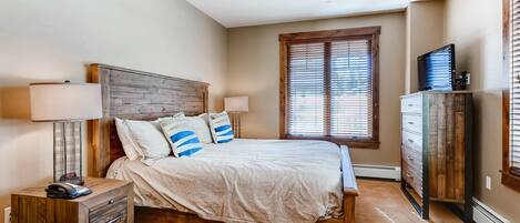 A bedroom with a wooden bed, two bedside lamps, a nightstand, a dresser, and a window with blinds.
