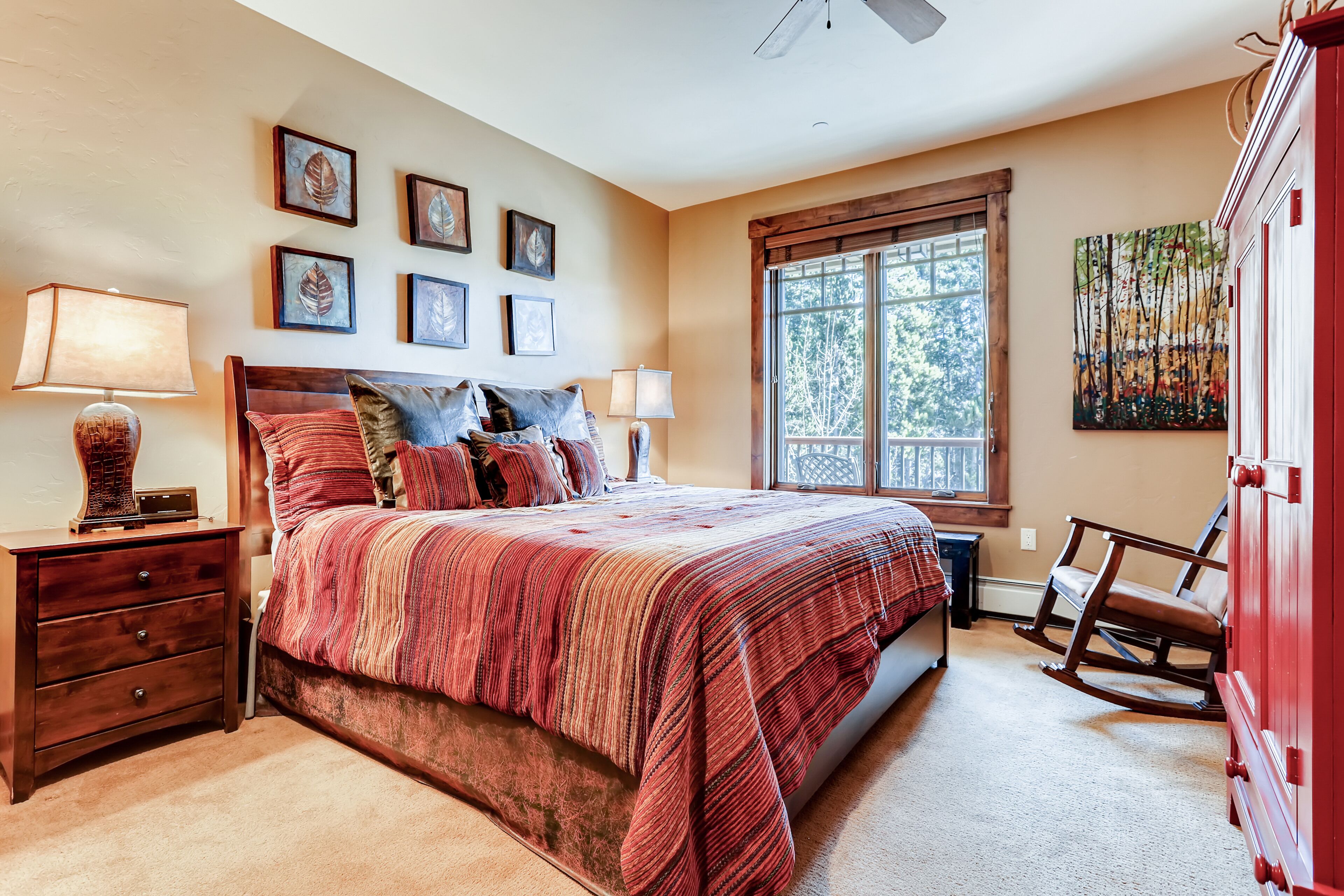 A bedroom with a large bed, two bedside tables, a rocking chair, and a window with a view of trees.
