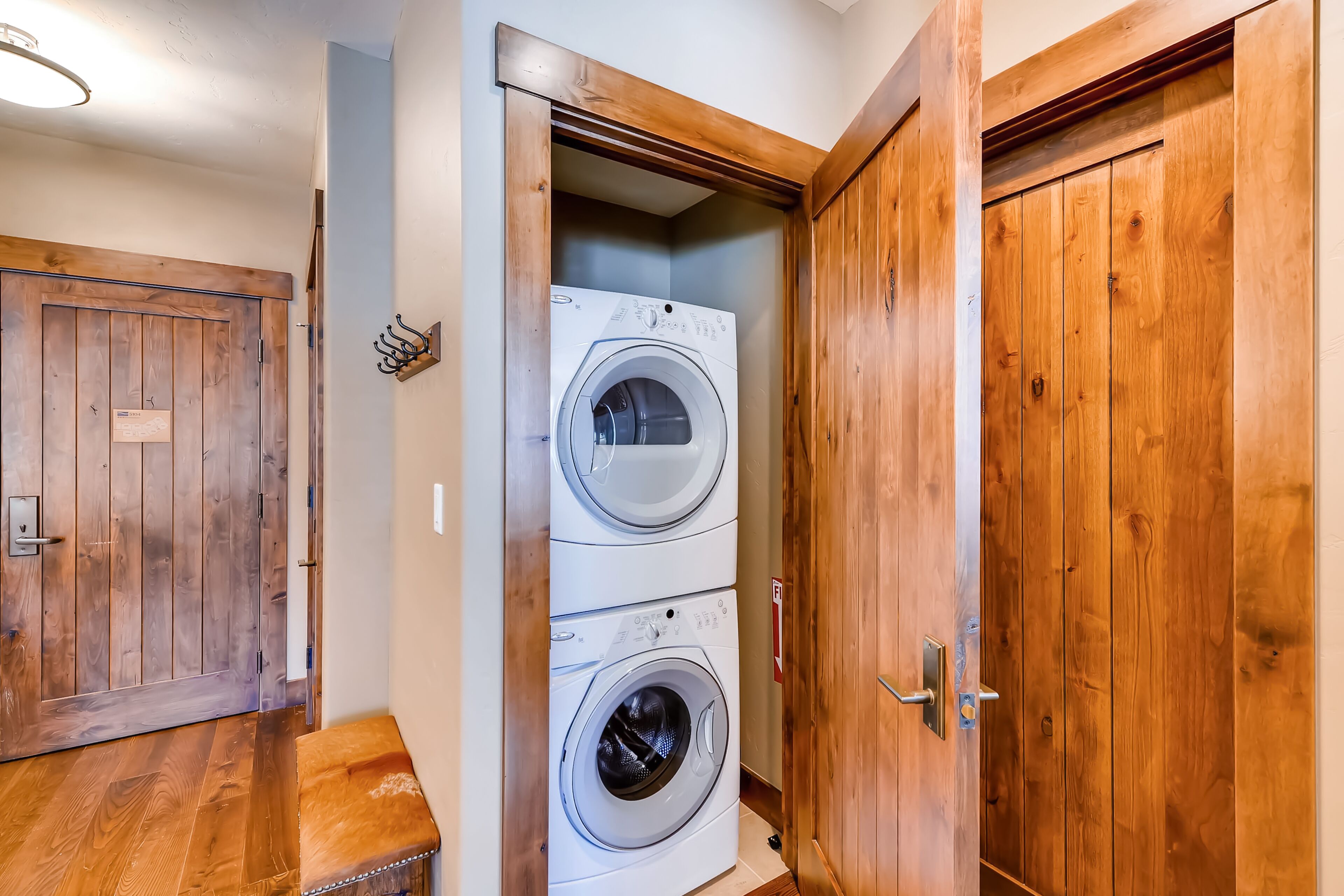 A laundry room with two stacked front-loading washing machines, wooden doors, and a bench.