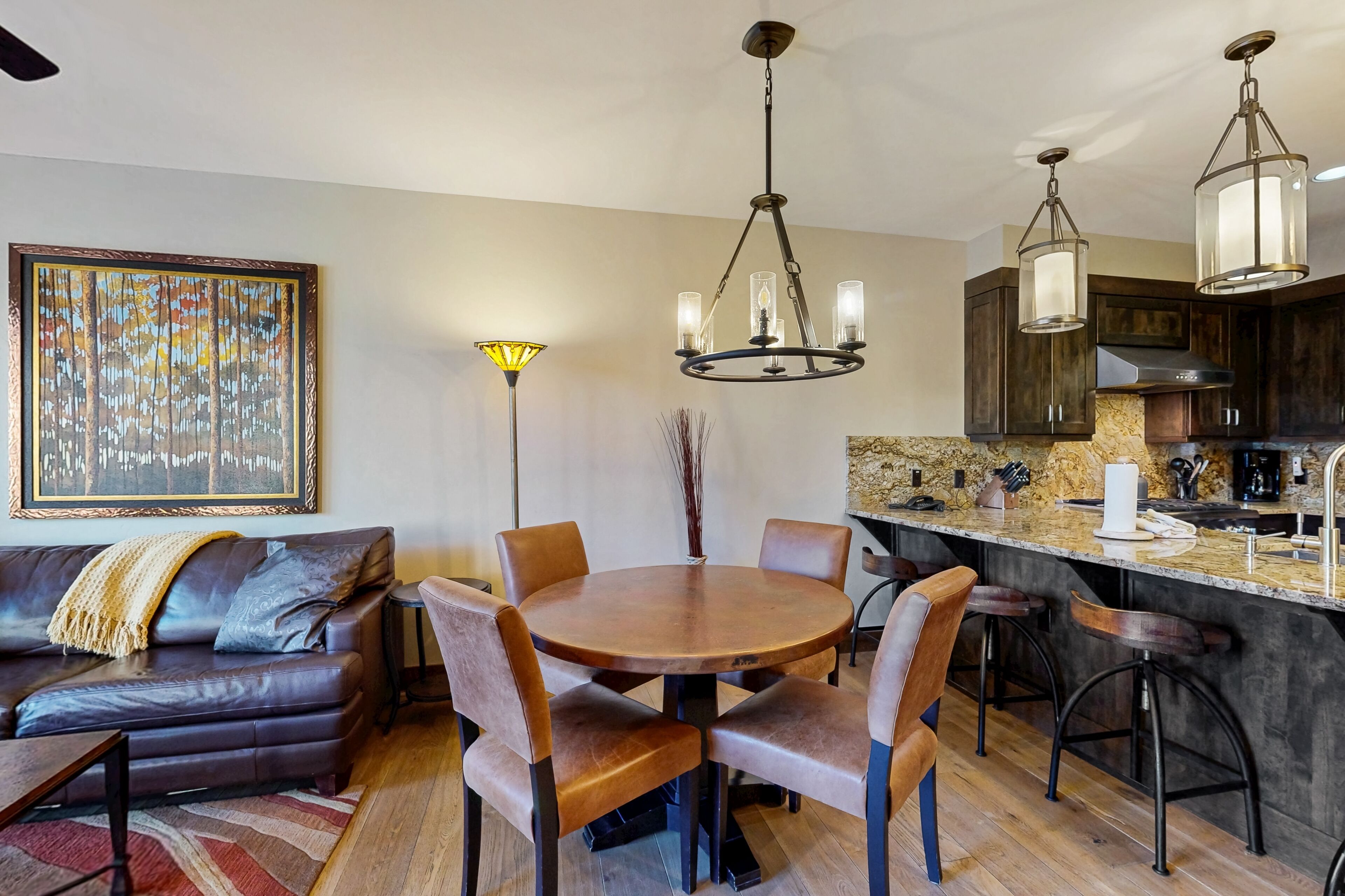 A modern living room with a dark leather sofa, a round wooden dining table with chairs, a chandelier, and a kitchen area with dark wooden cabinets and a marble countertop.