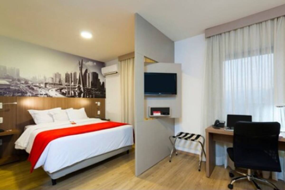 A hotel room with a bed, a desk, a chair, a TV, and a cityscape mural on the wall.