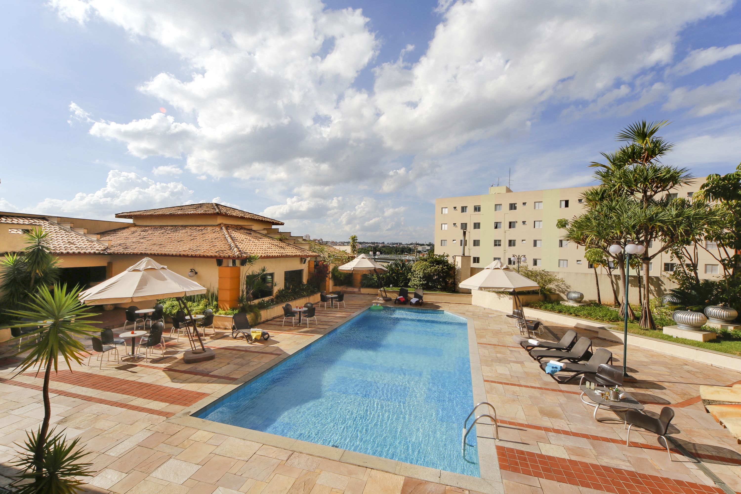 A swimming pool with lounge chairs and umbrellas, surrounded by buildings and palm trees.
