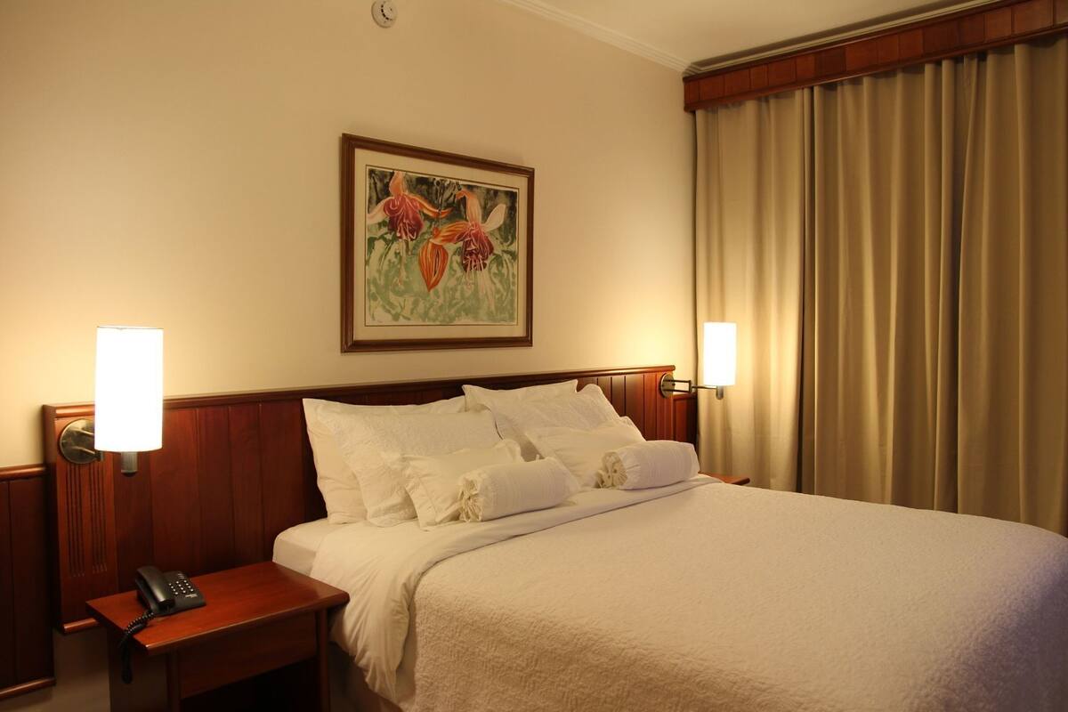 Une chambre d’hôtel avec un lit bien fait, un téléphone sur la table de chevet, un tableau au mur et une fenêtre avec des rideaux.