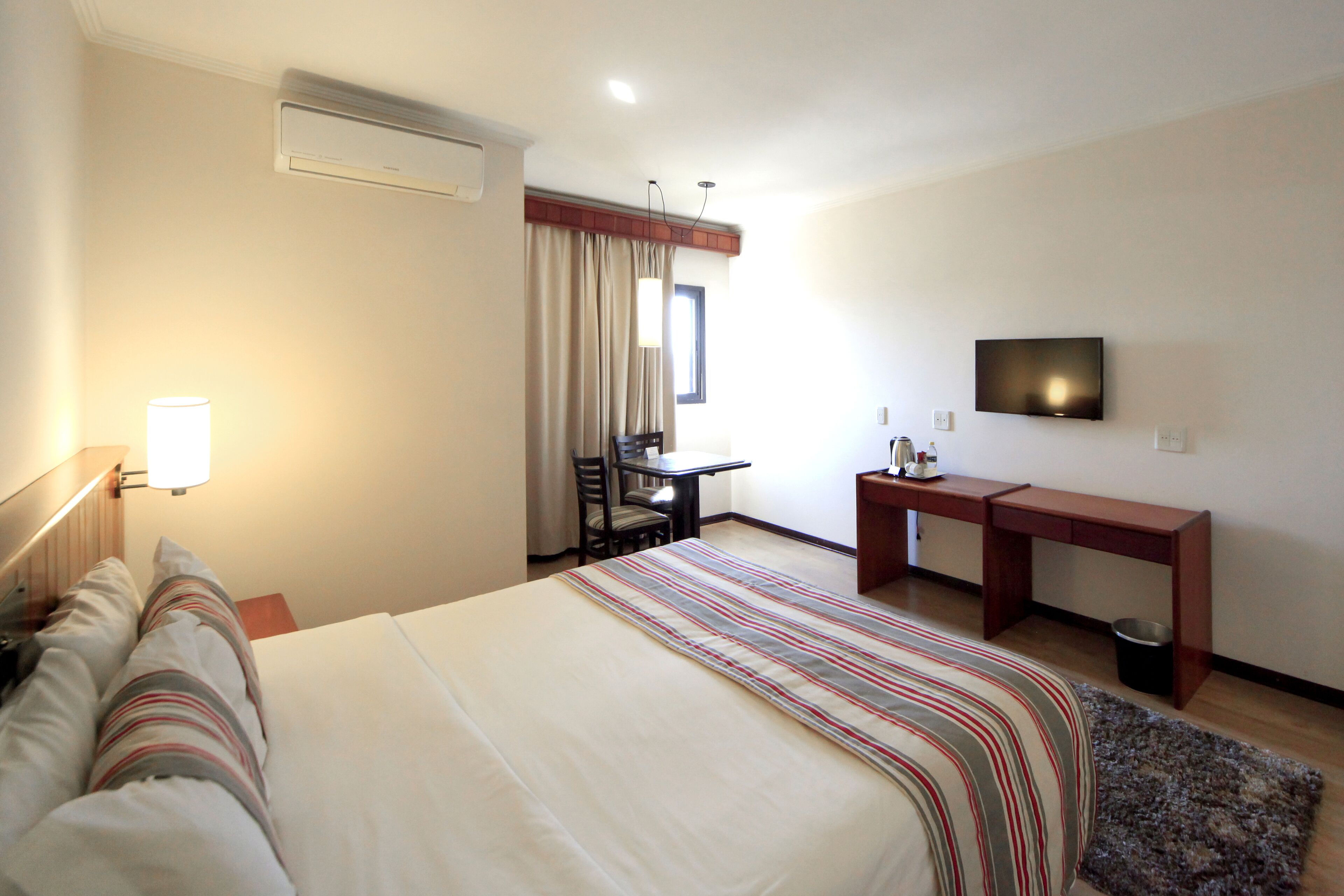 Una habitación de hotel con cama, escritorio, silla, televisor y ventana con cortinas.