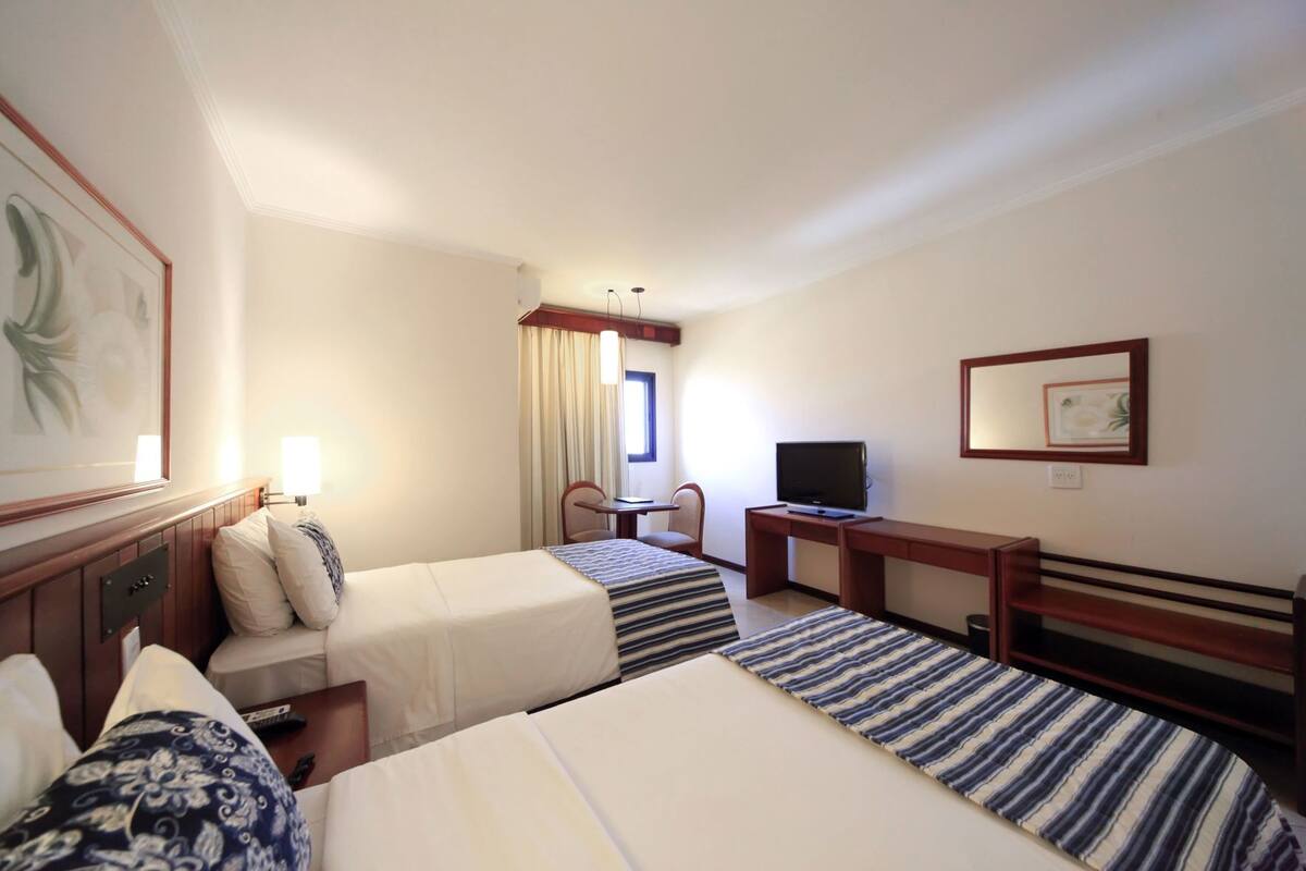 Une chambre d’hôtel avec deux lits, un bureau avec une télévision, une chaise et une fenêtre avec des rideaux.