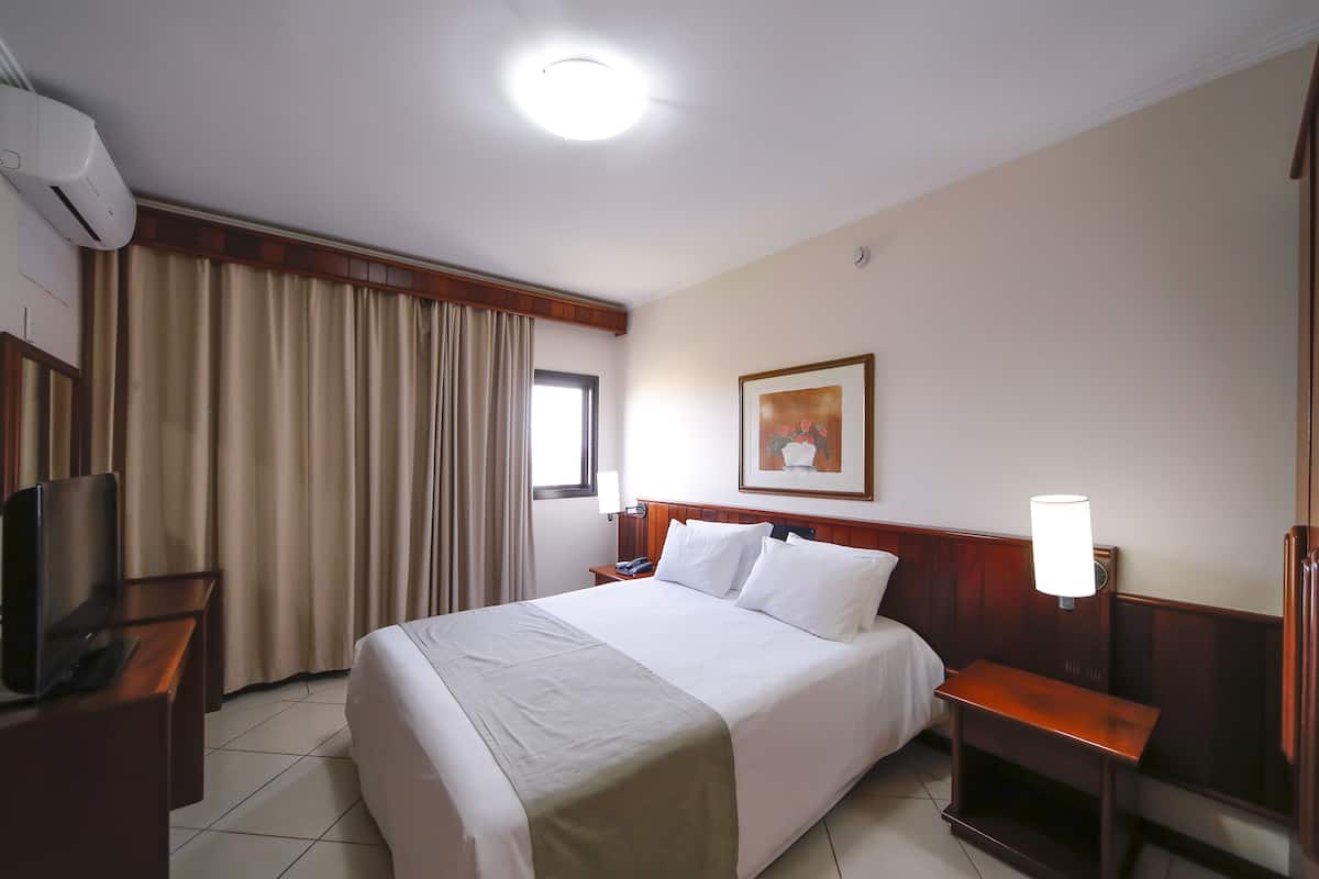 Une chambre d’hôtel avec un lit, un bureau avec une télévision, une table de chevet, une lampe et un tableau au mur.