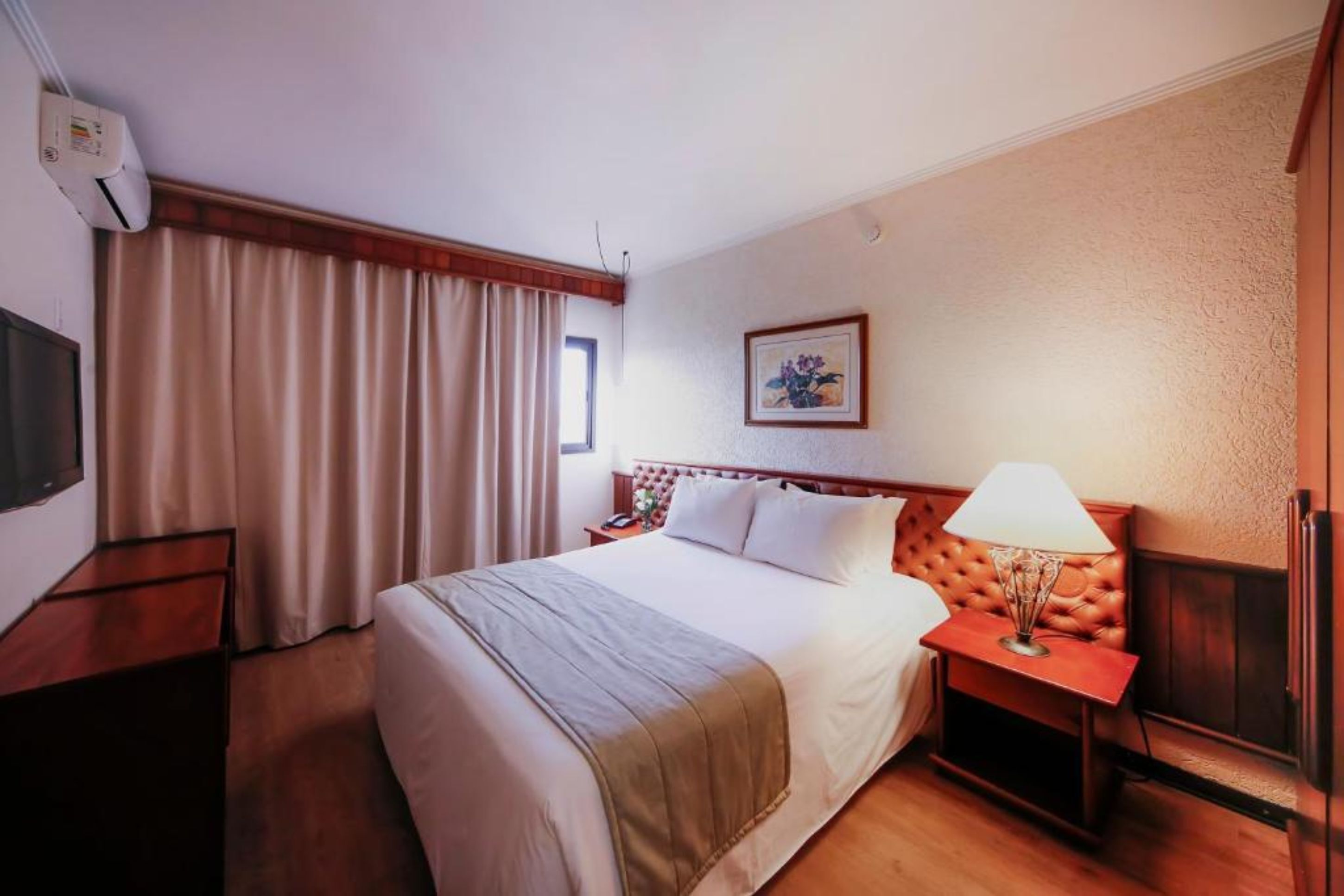 A hotel room with a bed, a nightstand, a lamp, a picture on the wall, and a window with curtains.