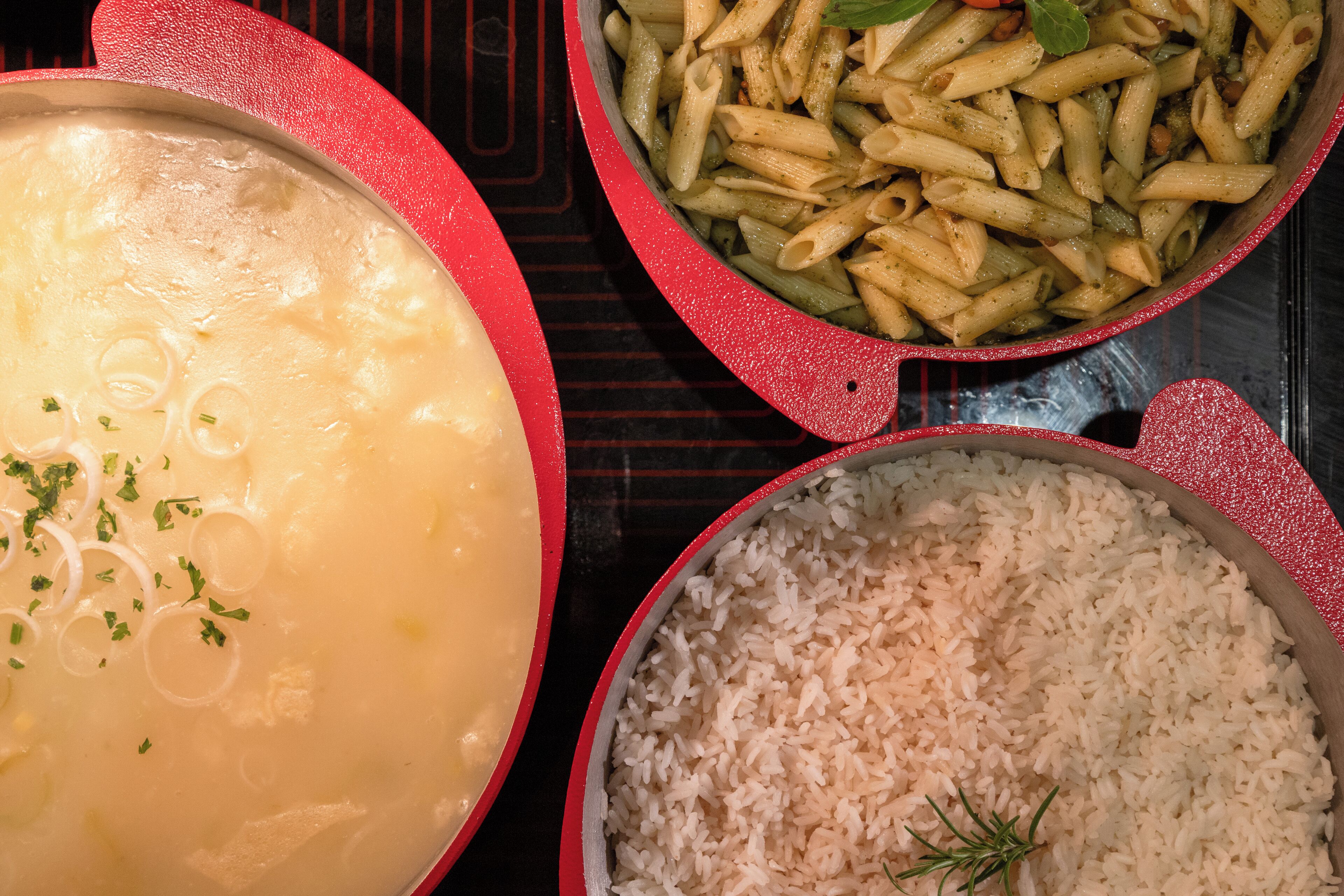 Une casserole rouge contenant une soupe crémeuse garnie d’herbes, une autre contenant des pâtes cuites et une troisième contenant du riz cuit.