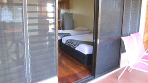 Een kamer met een schuifdeur naar een balkon, een bed met een gebedrukte sprei, een klein kitchenette en een roze stoel.
