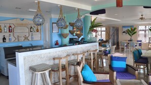 Amplia zona de bar con mostrador y varios taburetes, rodeada de mobiliario de mimbre e iluminación decorativa colgante.