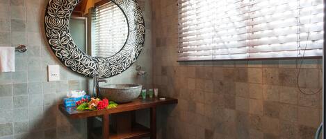A bathroom with a round mirror, a sink with toiletries, a trash bin, and a woven stool.