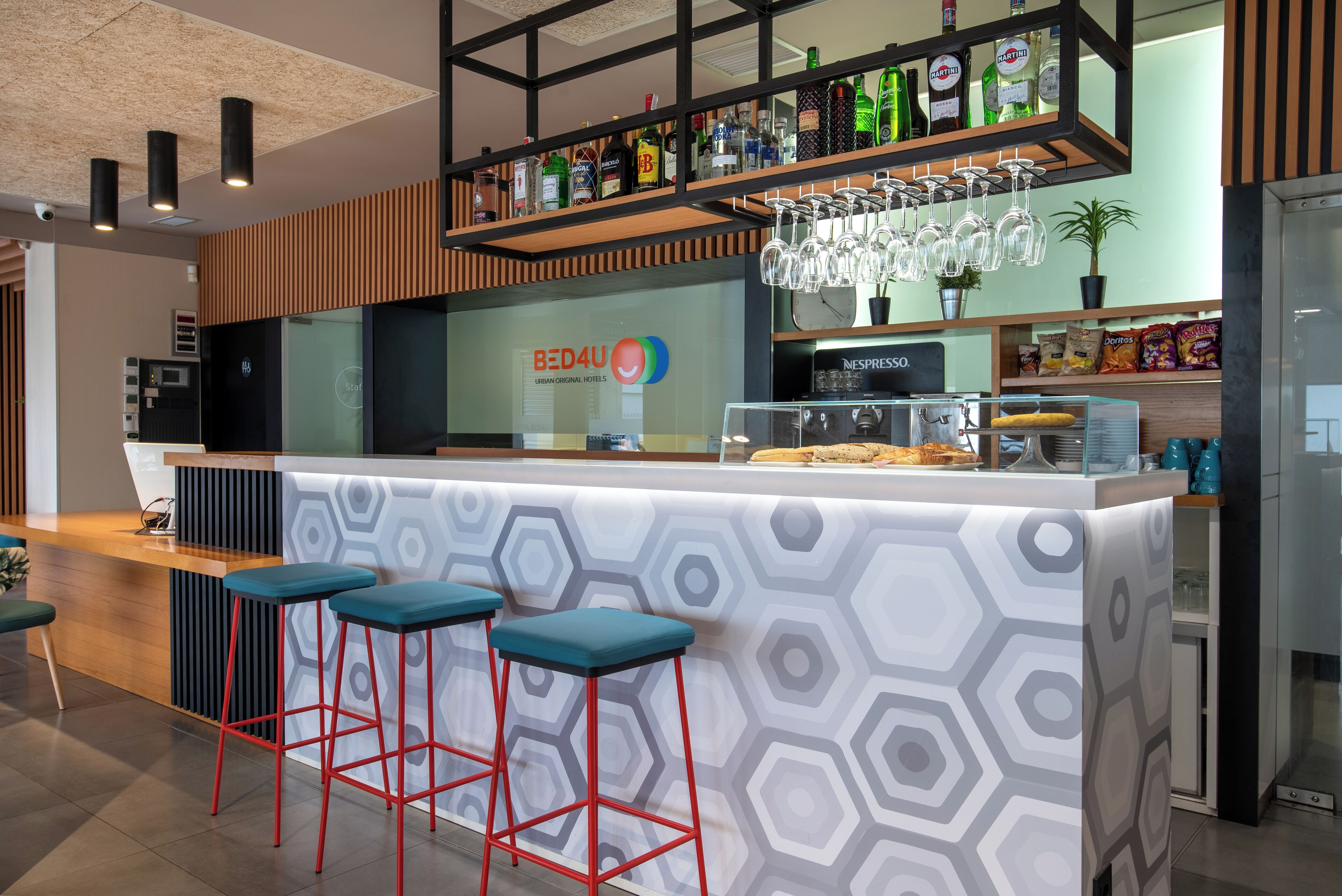 Een moderne barruimte met een wand in een honingraatpatroon, rode barstoelen en diverse drankjes op planken.