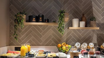 A breakfast buffet with various food items and beverages, including fruits, pastries, and condiments, displayed on a counter with hanging plants and shelves above.