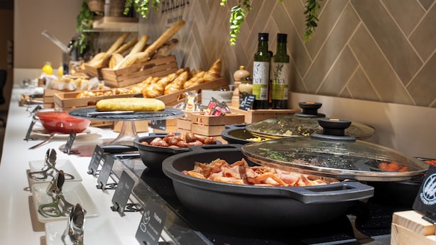 Un bufé con diversos alimentos, entre ellos pan, bollería y carnes, dispuesto en una barra con una pared de azulejos y plantas colgantes al fondo.