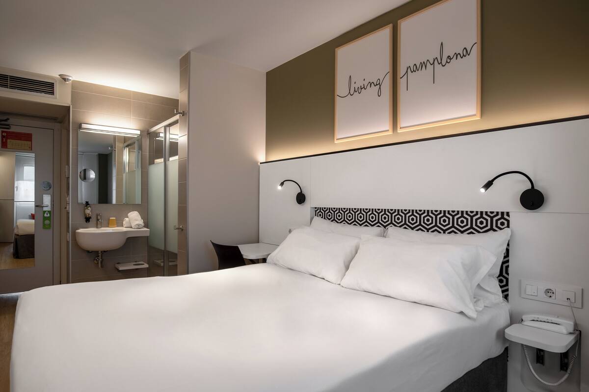 Une chambre d’hôtel avec un grand lit, une table de chevet et un miroir. Un panneau encadré est accroché au mur, sur lequel on peut lire : « Living Pamplona ».