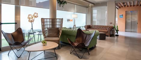 A modern hotel lobby with a green sofa, brown leather chairs, a round table, and a large window offering a view.