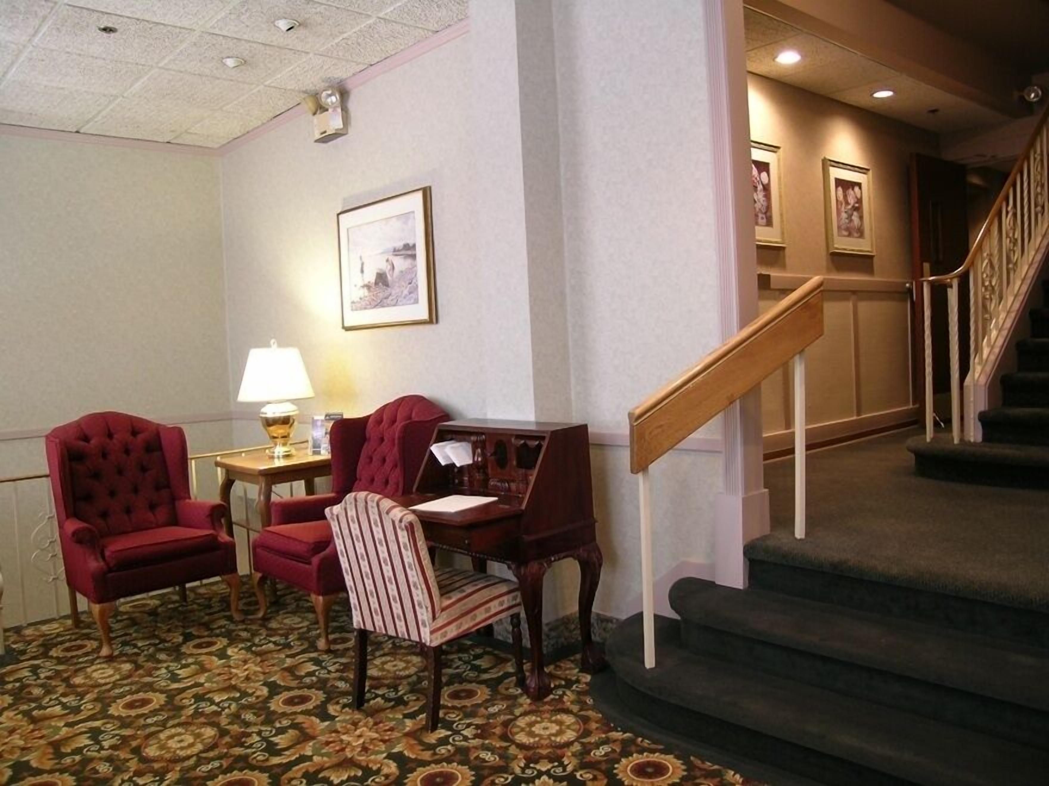 A hotel lobby with a carpeted floor, two armchairs, a small table with a lamp, a framed picture on the wall, and a staircase with a wooden handrail.