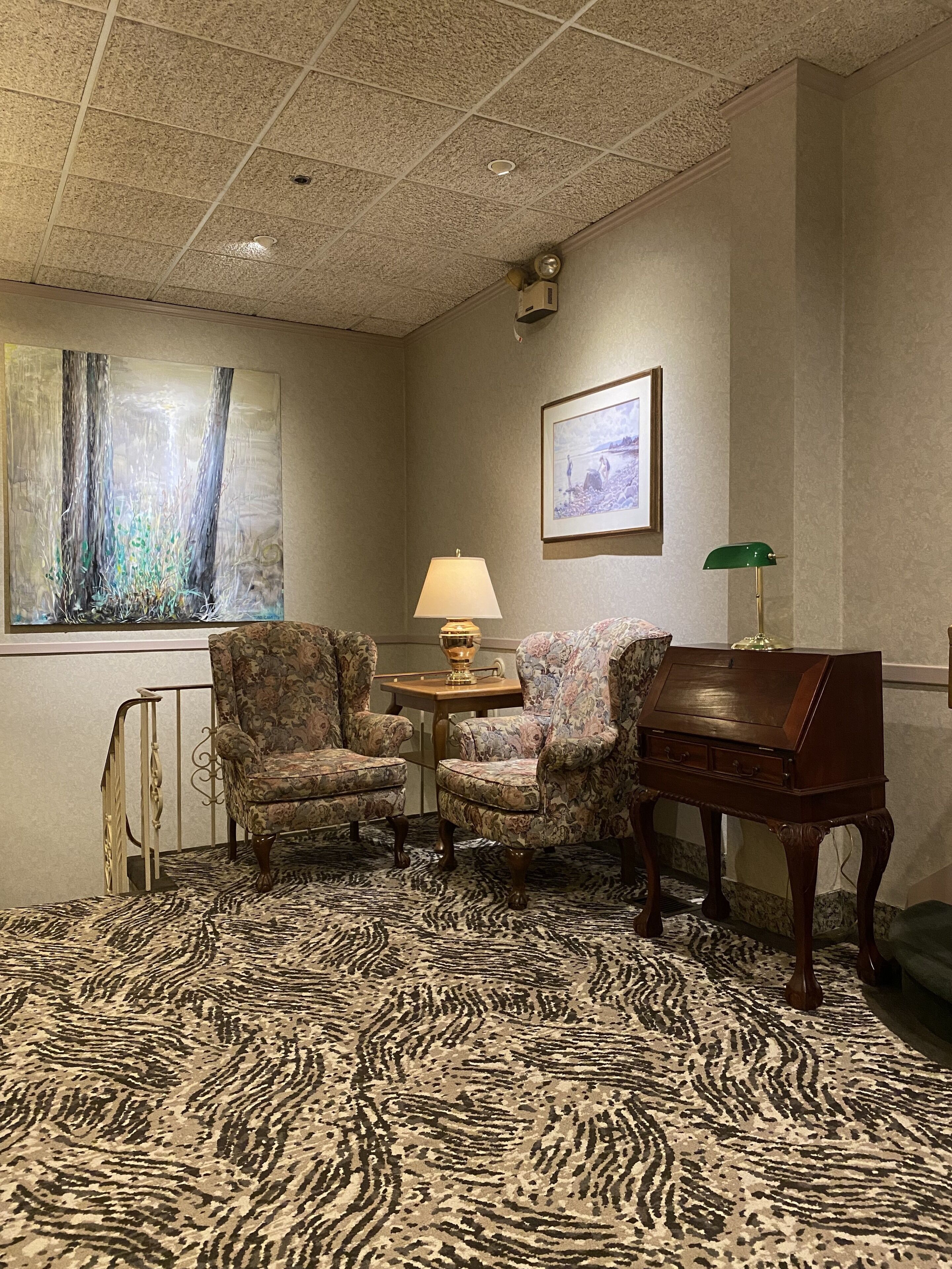 A room with a zebra-patterned carpet, two floral armchairs, a wooden sideboard, a table with a lamp, and a framed picture on the wall.