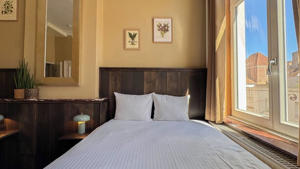 A hotel room with a large bed, a nightstand with a lamp, a mirror, and two framed botanical prints on the wall. - Botaniek (Bruges)