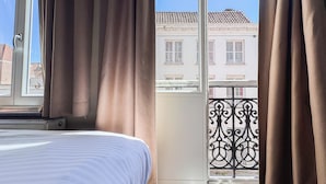 A hotel room with a bed, curtains, a view of buildings, and a balcony with a wrought iron railing. - Botaniek (Bruges)