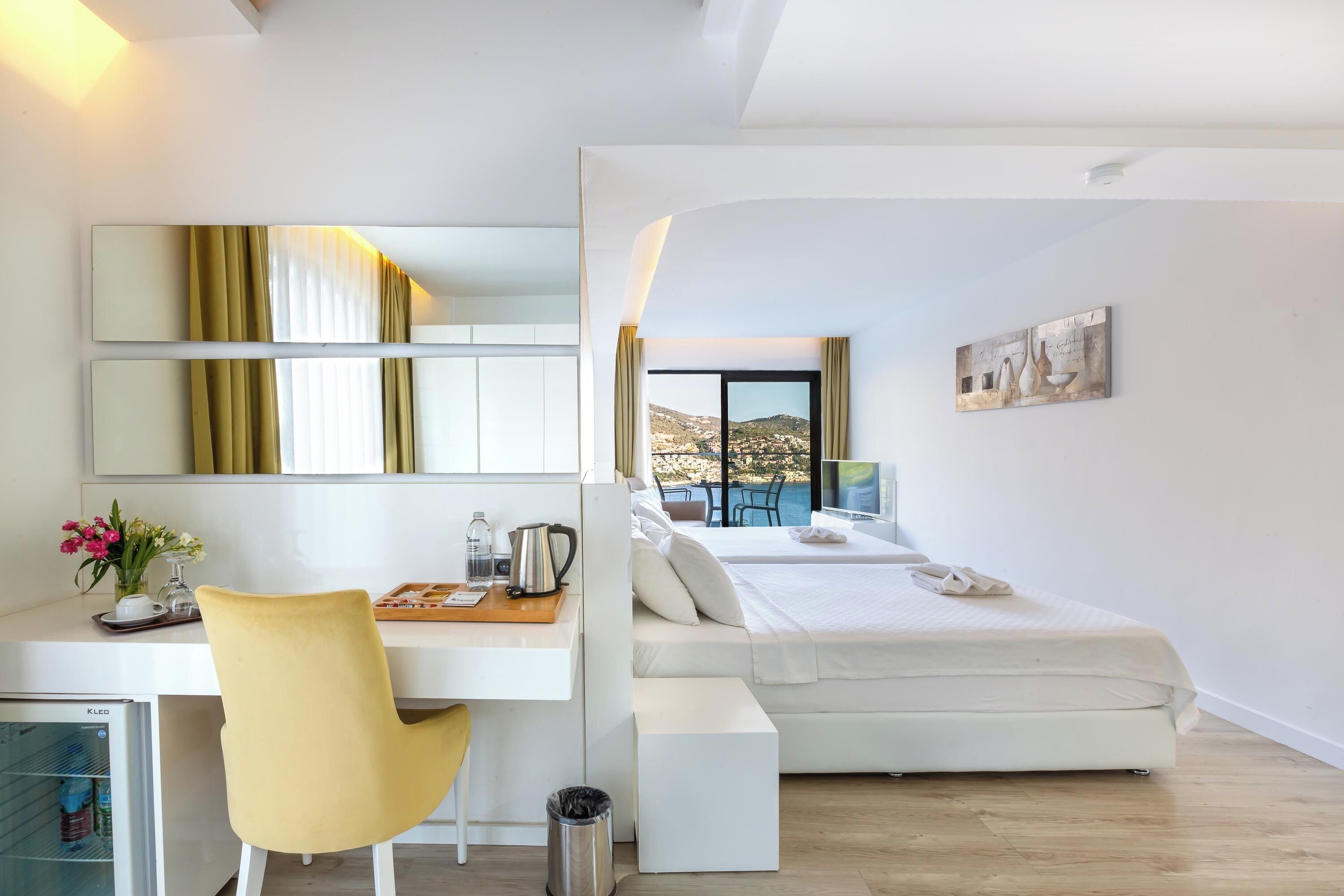 Une chambre d’hôtel moderne avec un bureau blanc, une chaise jaune, un lit donnant sur la ville et un petit réfrigérateur.