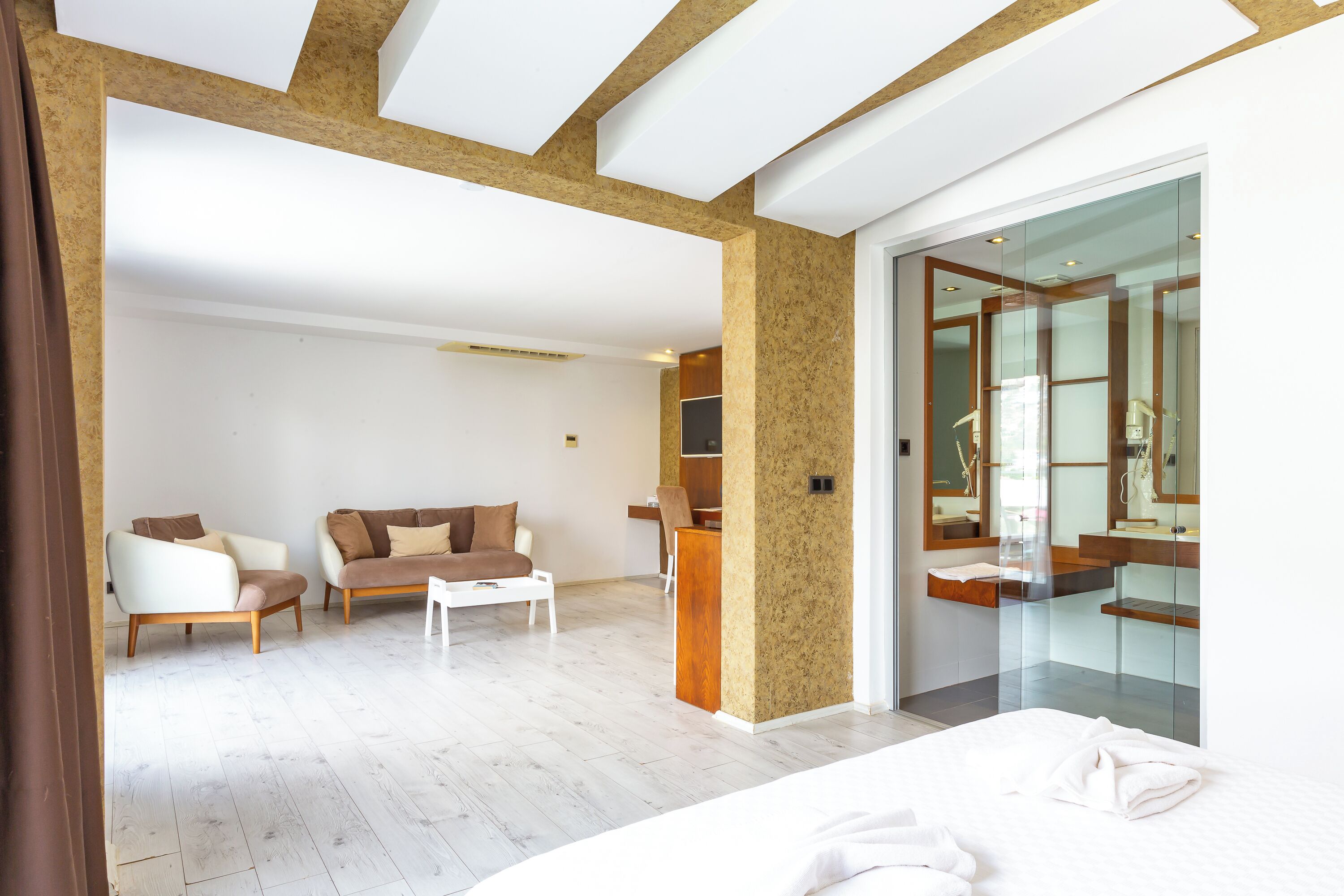 A modern hotel room with a large bed, a sitting area with a sofa and armchairs, a small table, and a glass-enclosed bathroom with a wooden vanity.