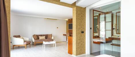 A modern hotel room with a large bed, a sitting area with a sofa and armchairs, a small table, and a glass-enclosed bathroom with a wooden vanity.