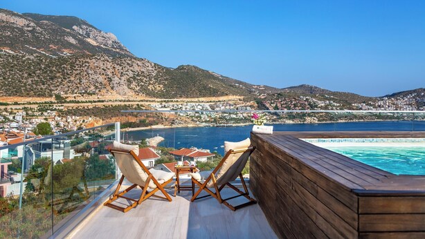 A wooden deck with two chairs and a table overlooking a coastal town and a swimming pool.