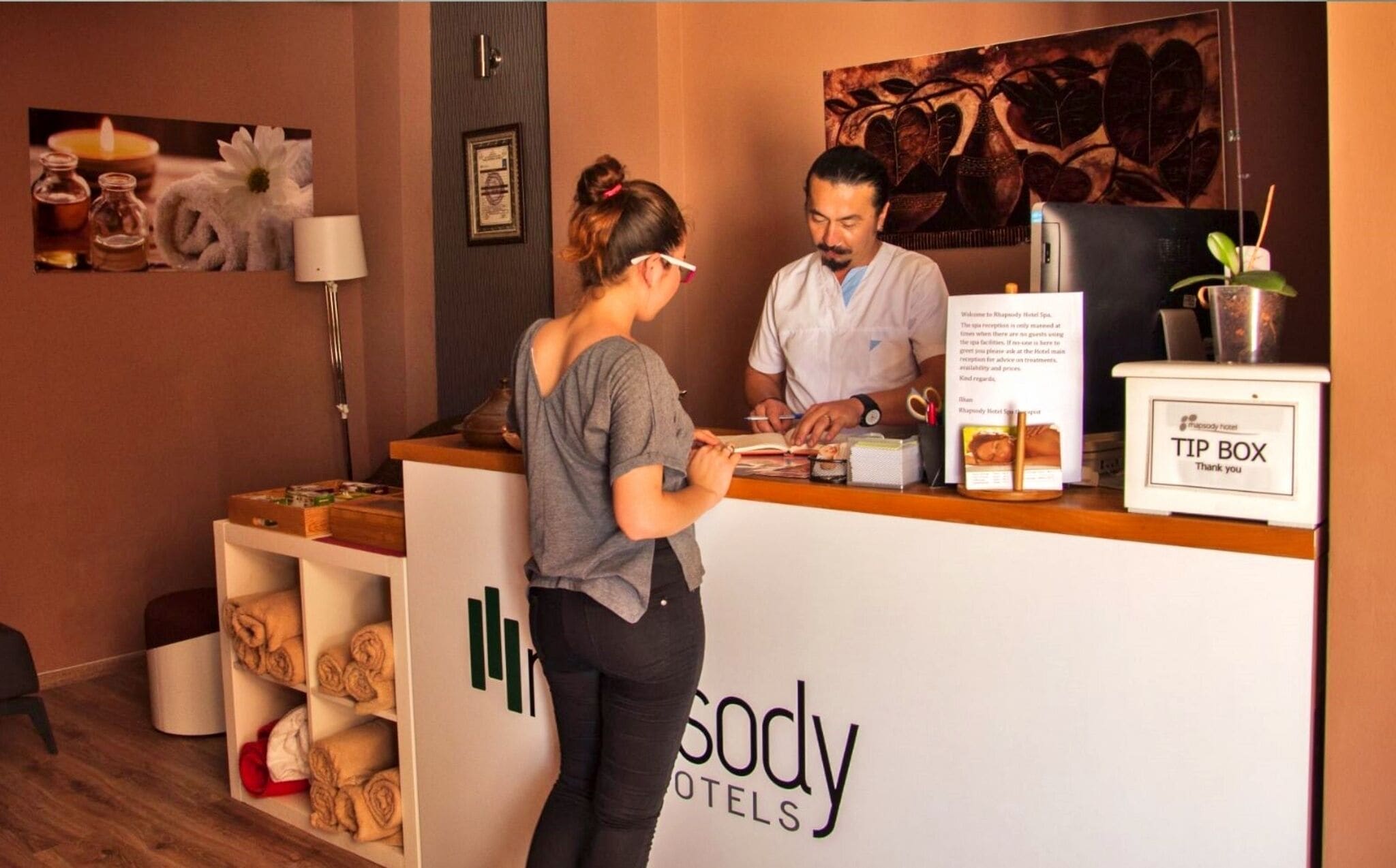 Un comptoir d’accueil dans un hôtel, avec une enseigne indiquant « Rhapsody Hotel Spa » et une autre indiquant « pourboires ». On y trouve des serviettes et une plante.
