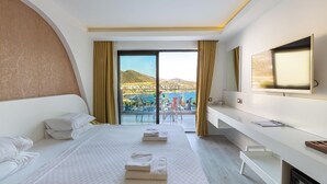 A hotel room with a bed, a TV, a desk, and a balcony with a view of the sea and mountains.