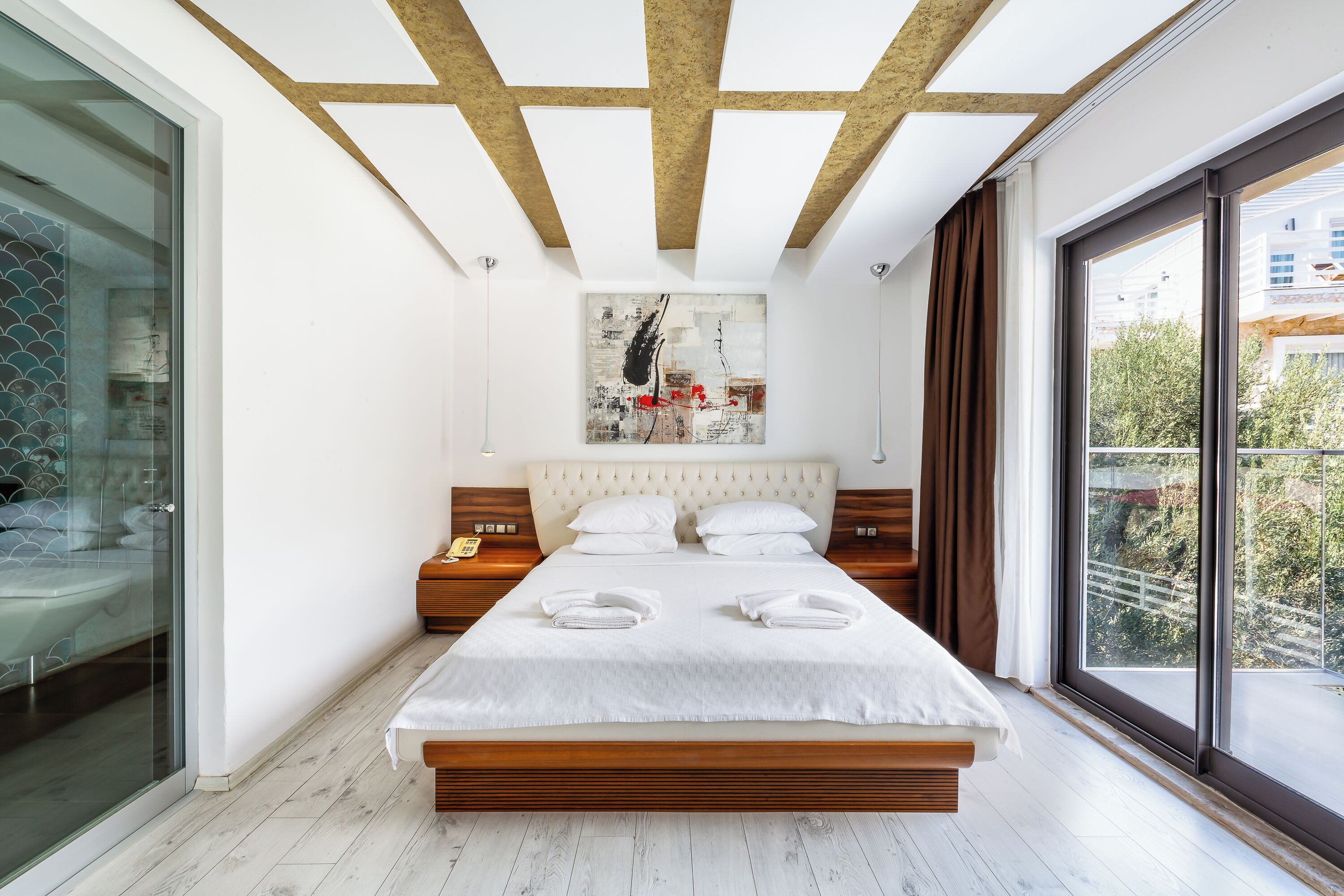 Une chambre moderne dotée d’un grand lit, d’une douche en verre, d’un plafond avec des poutres apparentes et offrant une vue sur la ville grâce à de grandes fenêtres.