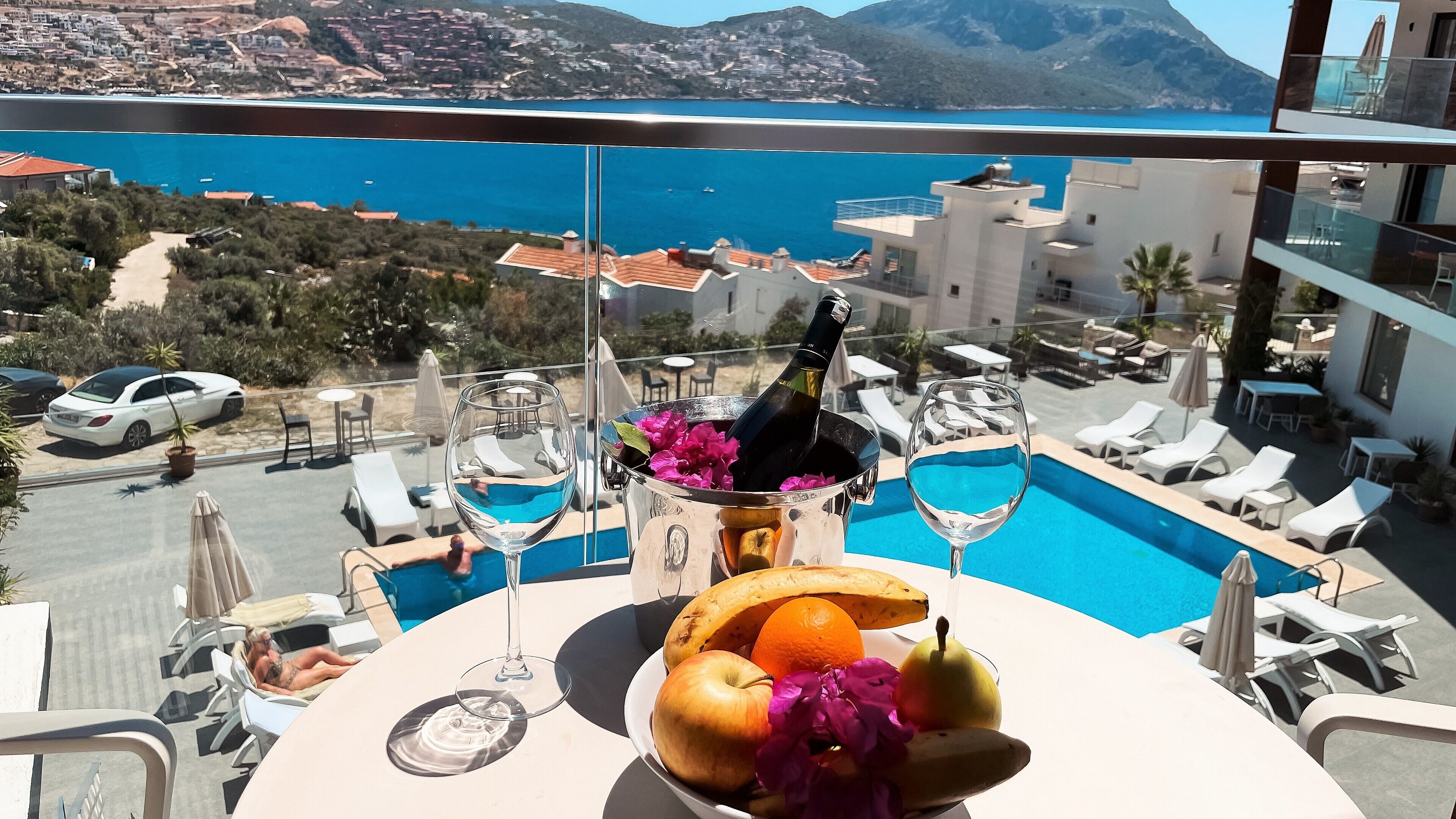 A dining table with a bottle of champagne, a glass, and fruit. A pool with lounge chairs is visible in the background.