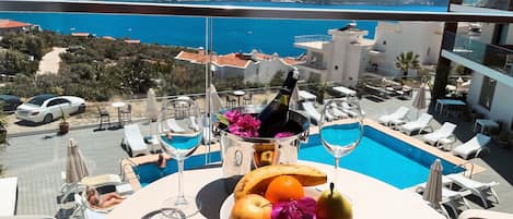 A dining table with a bottle of champagne, a glass, and fruit. A pool with lounge chairs is visible in the background.