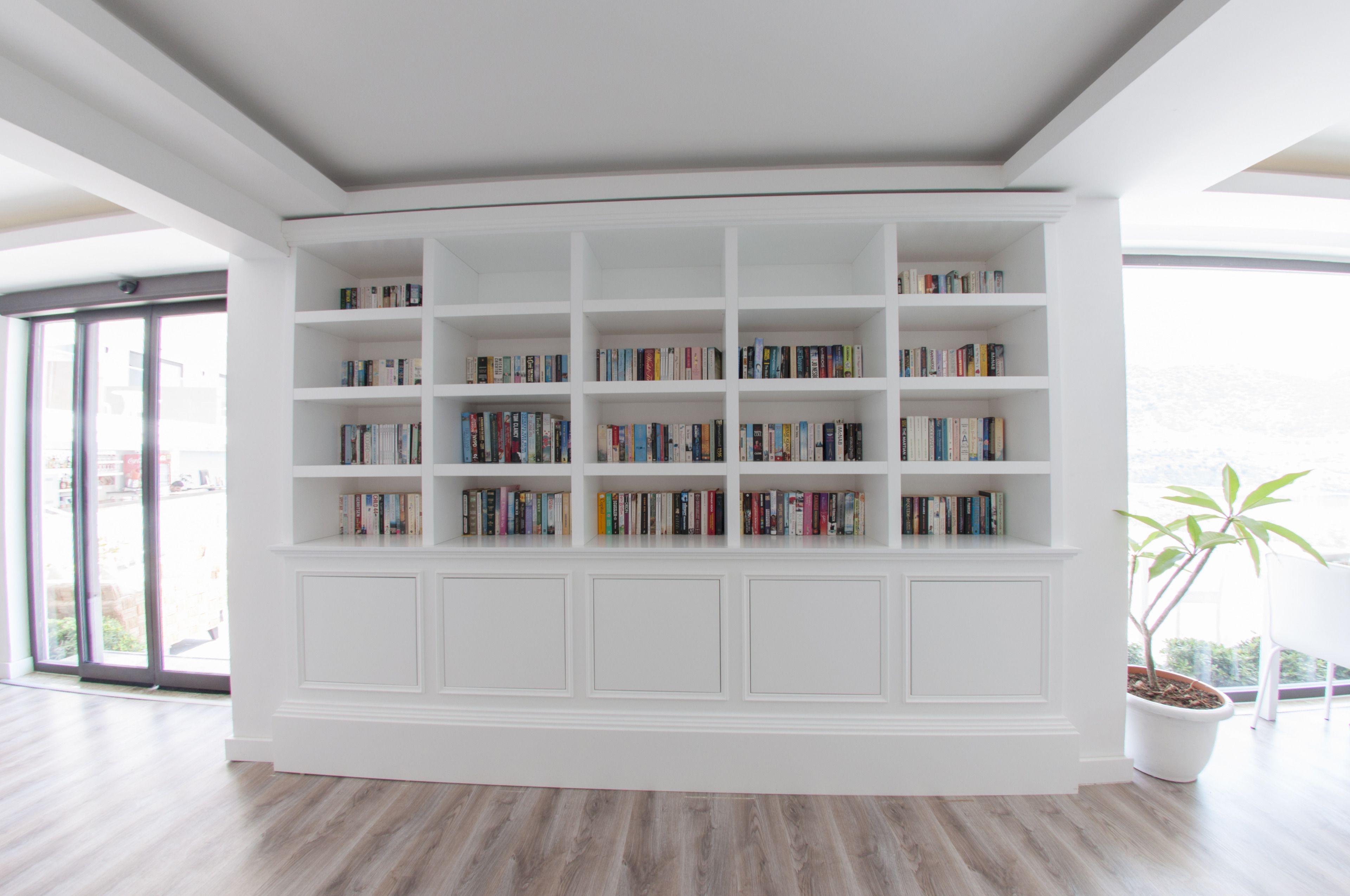 Ein modernes Wohnzimmer mit einem großen, weißen Einbauschrank voller Bücher, einer Zimmerpflanze und einem großen Fenster mit Blick ins Freie.