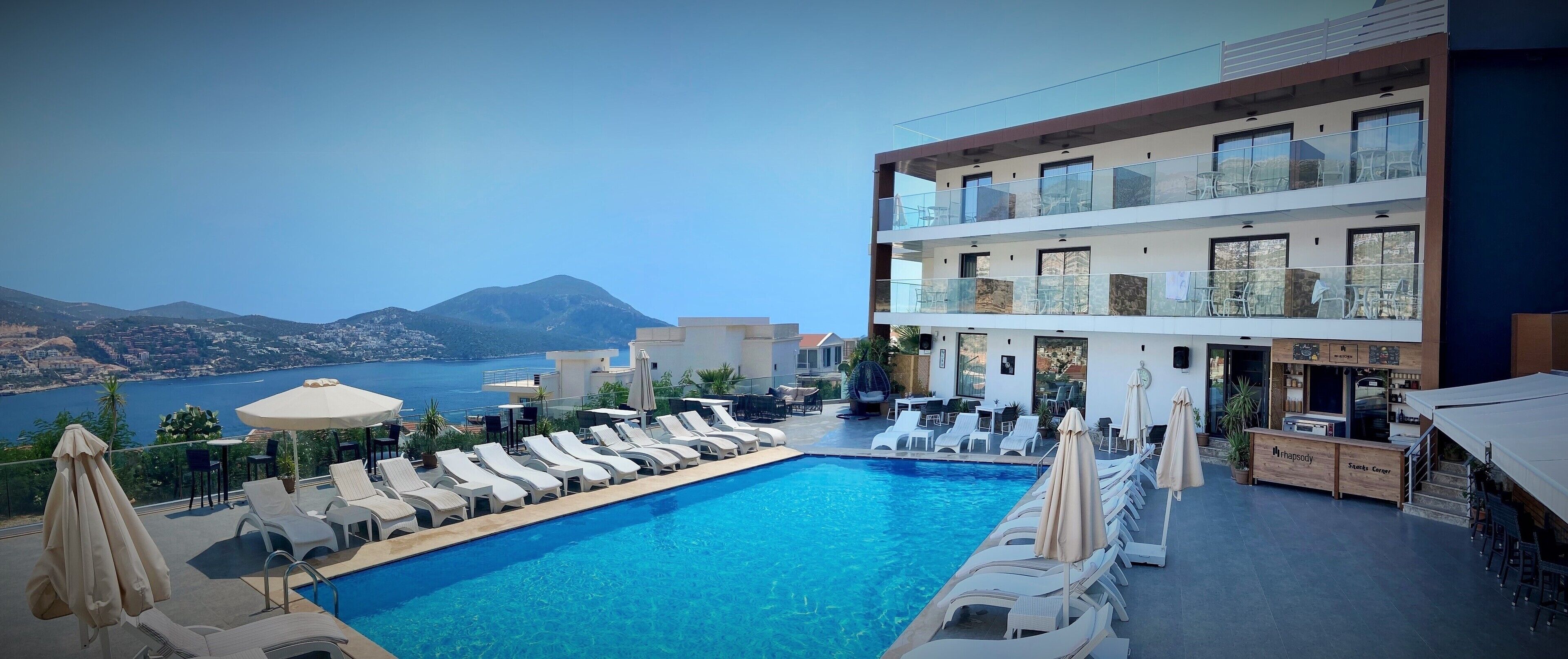 A rooftop pool with lounge chairs and umbrellas, overlooking a scenic view of a mountain and sea.