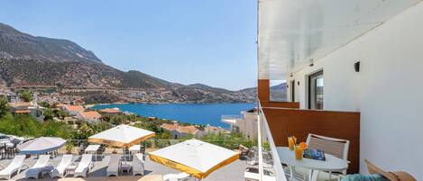 A balcony with a pool, lounge chairs, and umbrellas overlooking a coastal town and mountains.