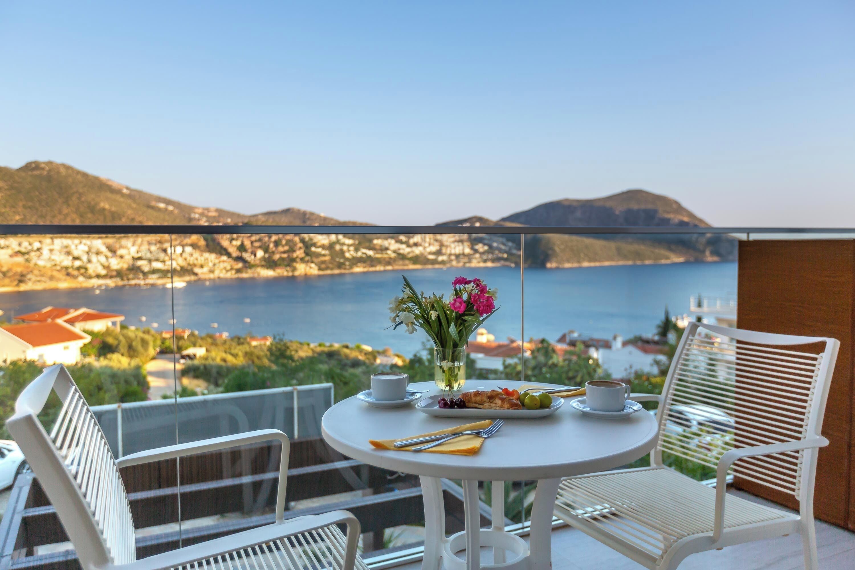 Un balcon avec une table dressée pour deux personnes, un vase rempli de fleurs et une vue imprenable sur une ville côtière.