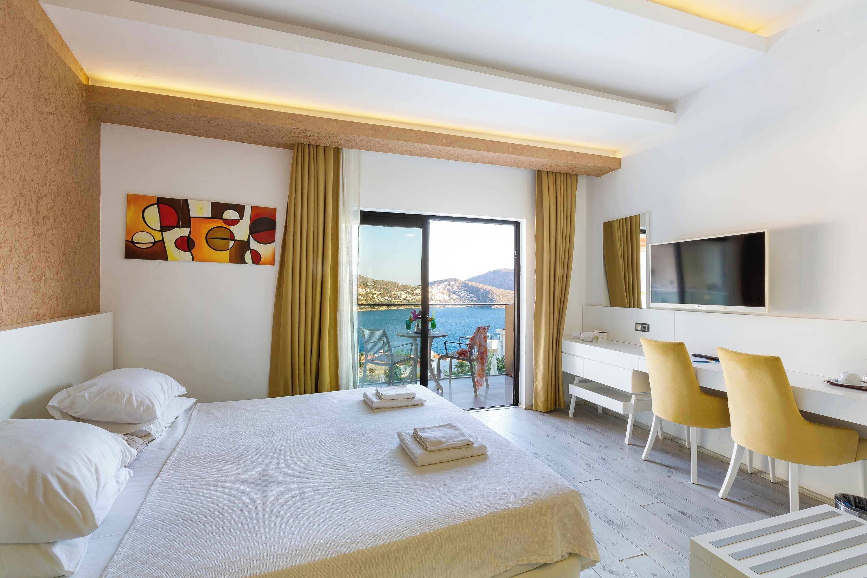 Habitación de hotel con cama, escritorio con sillas, televisor y balcón con vista al mar.