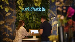 A reception desk with a digital screen displaying a message, surrounded by lush green foliage and palm leaf patterns on the wall. - Auberge du Bonheur (Tilburg)