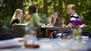 A group of people seated at an outdoor table with wicker chairs, surrounded by greenery and a lamp post. - Auberge du Bonheur (Tilburg)