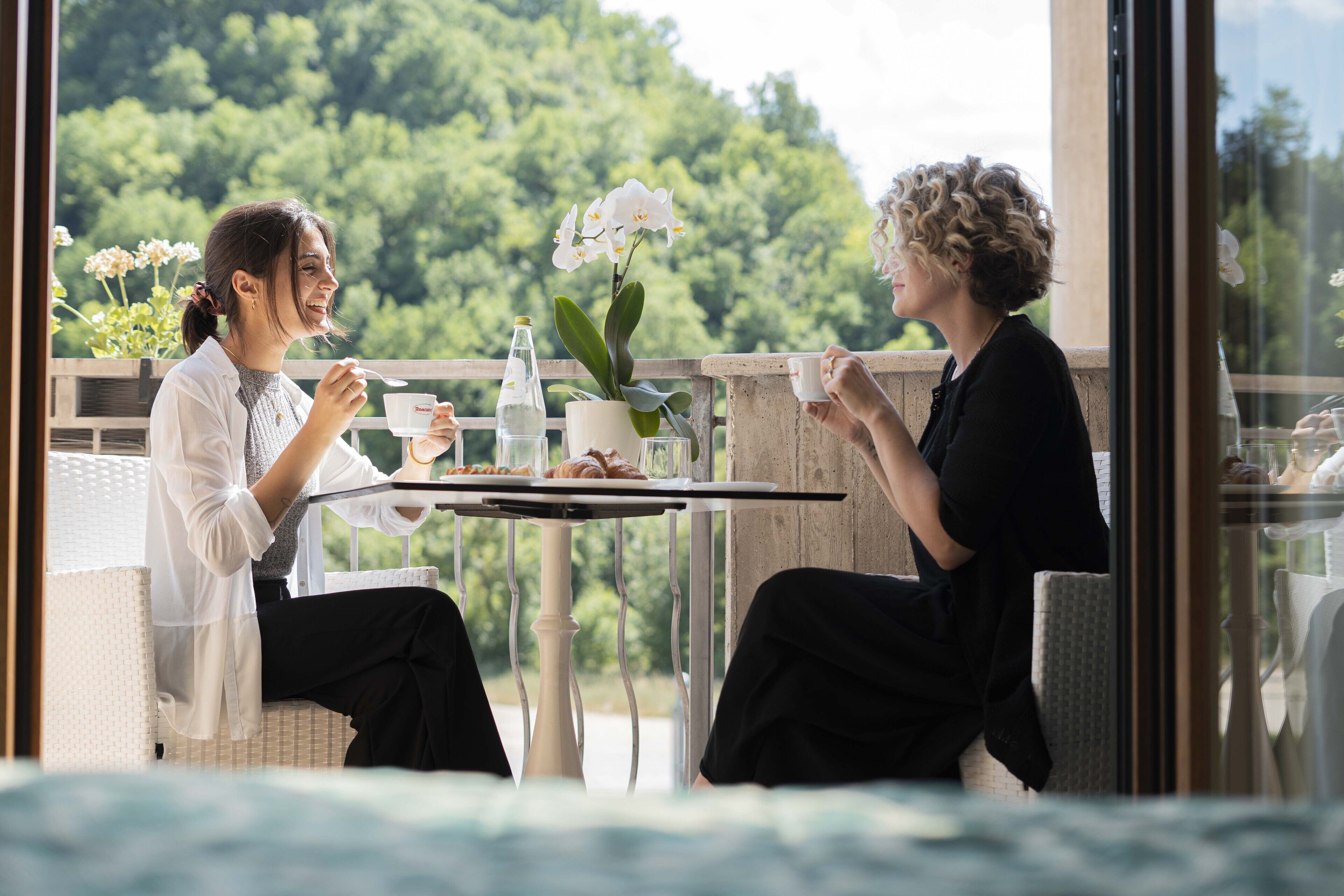 Deux personnes sont assises à une table, devant laquelle on aperçoit de la verdure, une plante en pot et une bouteille.