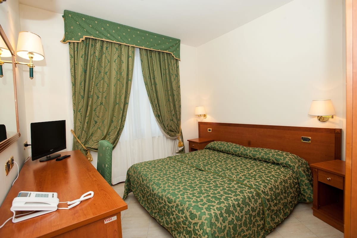 A hotel room with a bed, a desk with a telephone, a television, and two wall-mounted lamps.