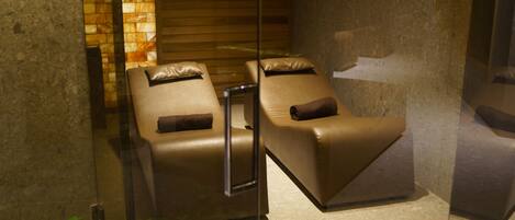 A spa room with two massage chairs, a glass partition, and a stone wall with a wooden panel.