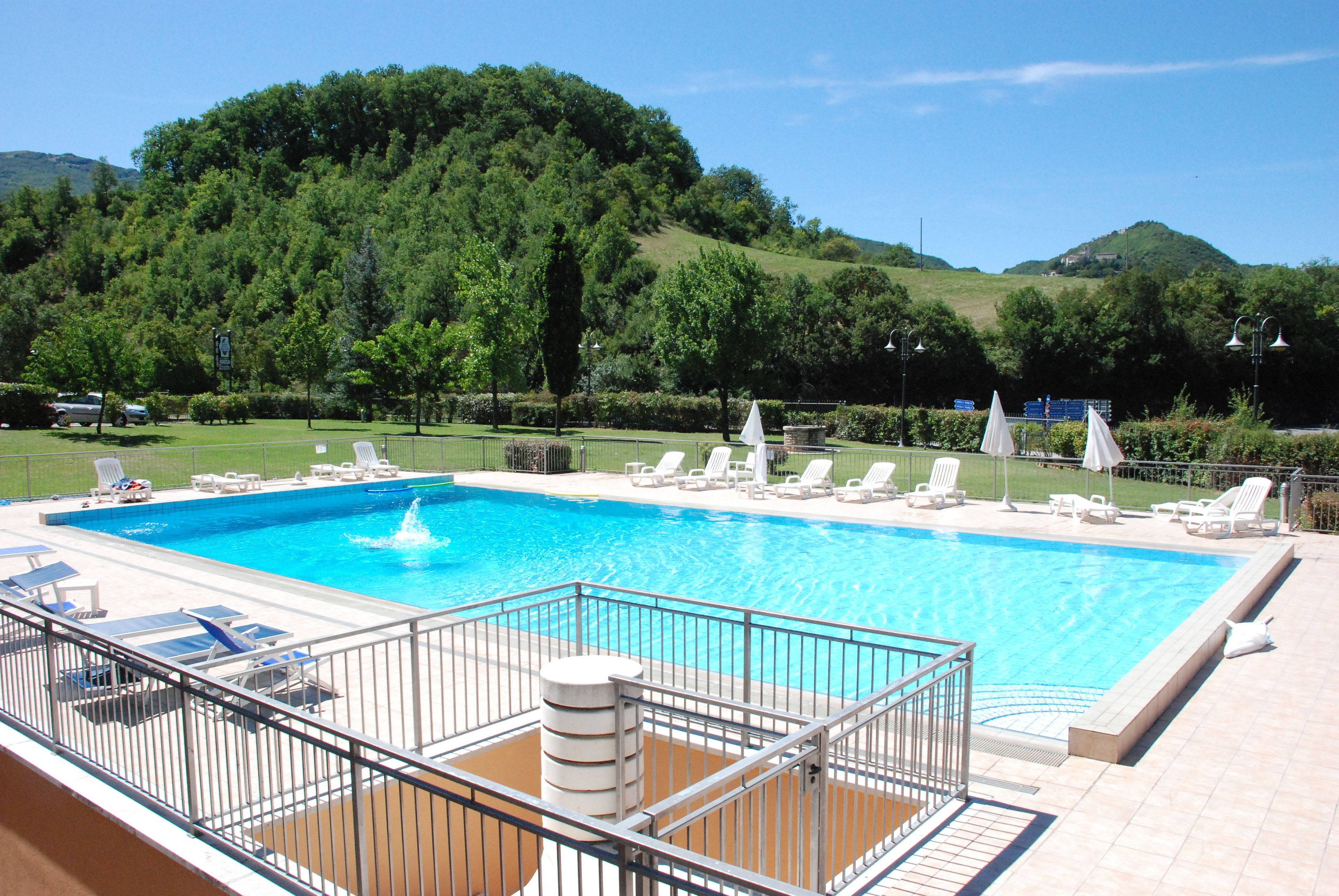 A rectangular swimming pool with a fountain, surrounded by lounge chairs and umbrellas, set against a backdrop of lush green hills.