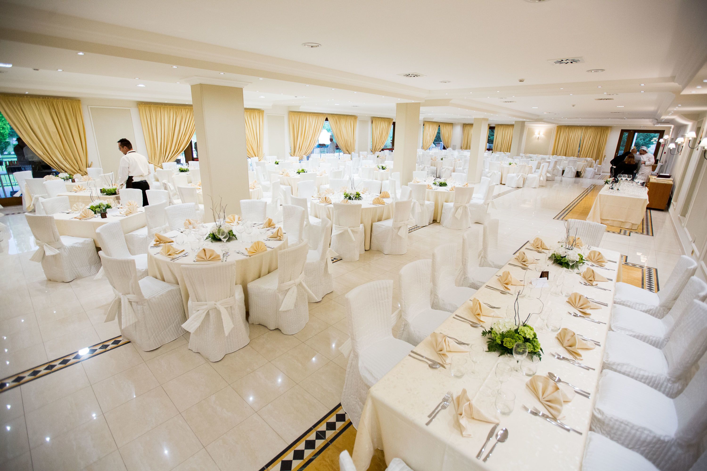 A large banquet hall with round tables set for dining, white chairs, and gold drapes.