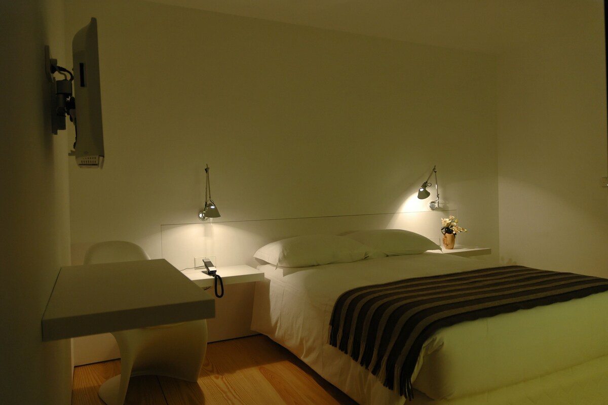 A hotel room with a bed, bedside table, lamp, and a desk with a chair.