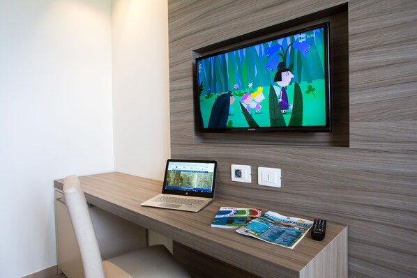 A hotel room with a wooden desk, a laptop, a remote control, and a television displaying an animated show. - Hotel Village Club Santa Caterina (Scalea)